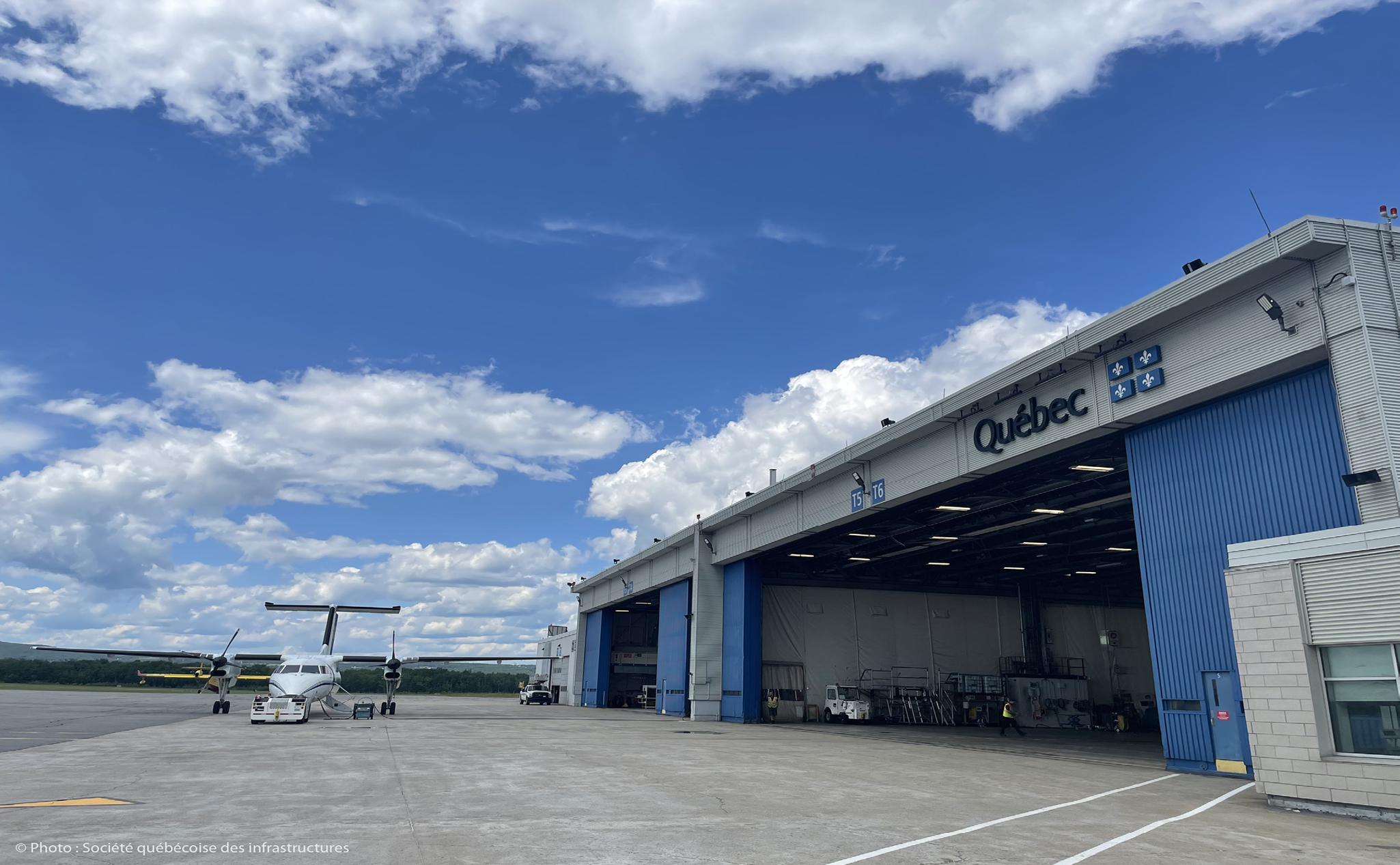 Un avion à hélices blanc est garé à l'extérieur d'un grand hangar bleu et blanc portant l'inscription "Québec", sous un ciel partiellement nuageux.