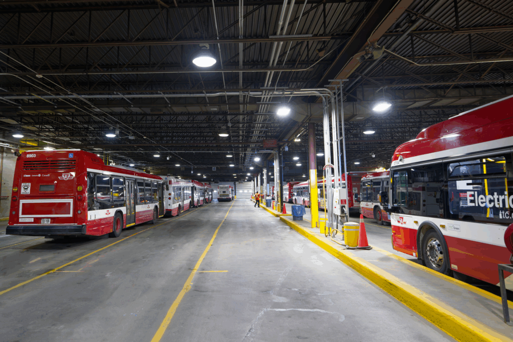 Several red and white city buses are parked inside a large, well-lit transit garage with high ceilings and exposed beams.