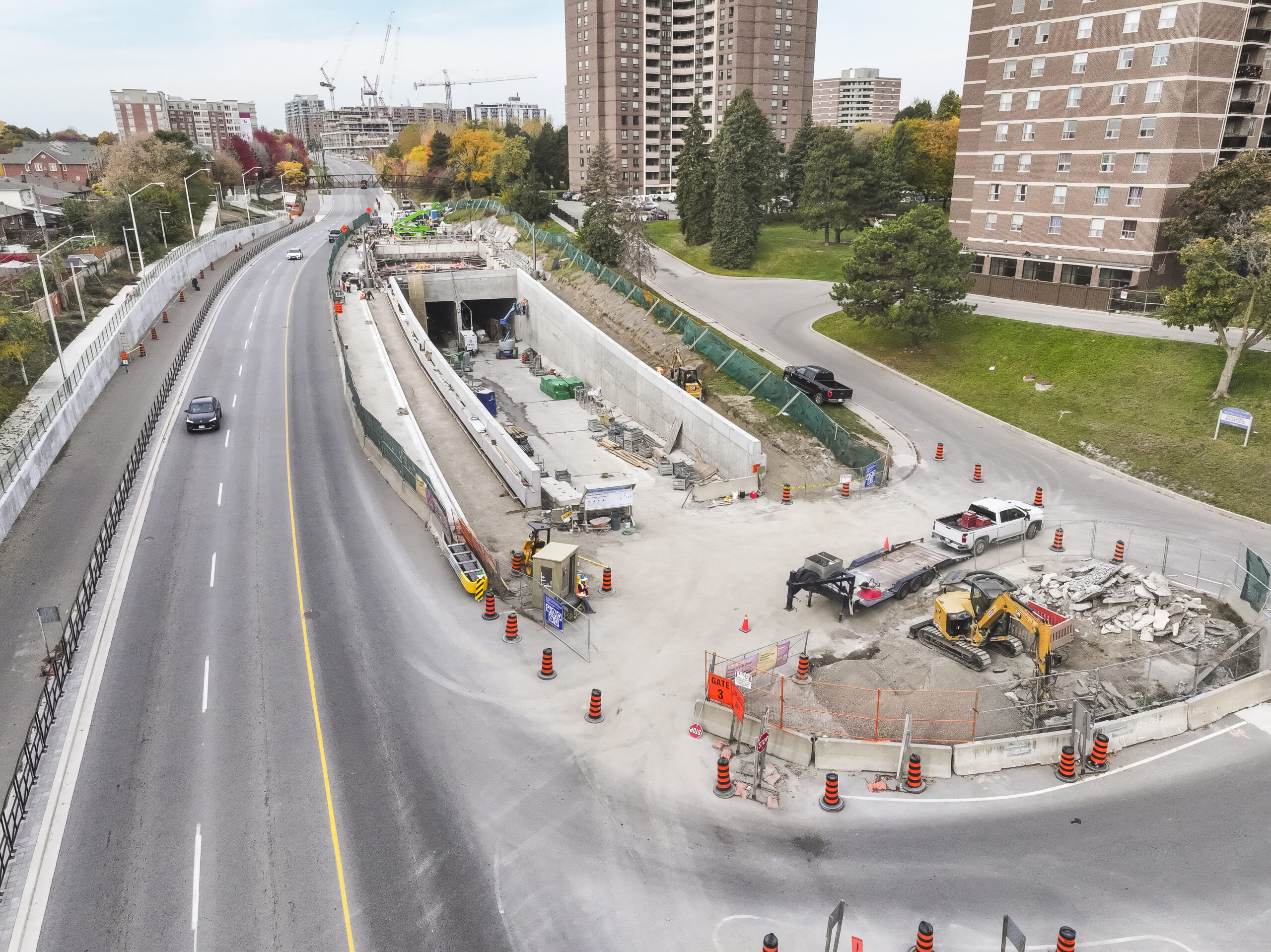 Aerial view of a construction site next to a busy road with several vehicles, construction equipment, and tall apartment buildings in the background.