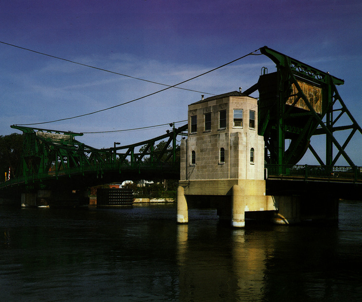 A green metal drawbridge spans a river, with a small concrete control house positioned on supports above the water.
