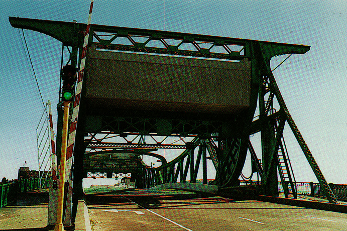 A green steel drawbridge is partially raised, with a red and white barrier lowered and a traffic light showing green on the left side.