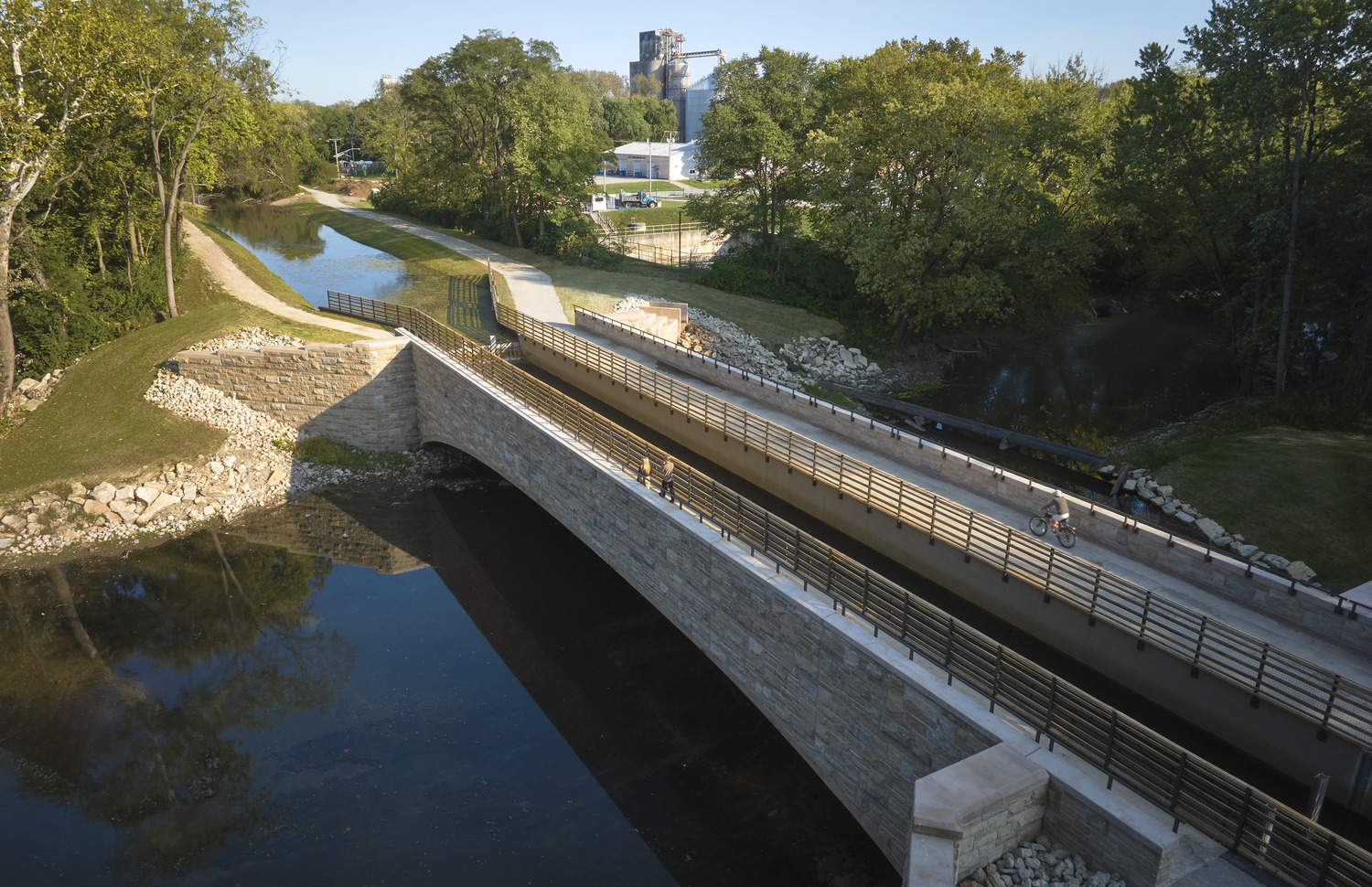 Aerial view of a modern stone bridge with bike and pedestrian paths crossing over a river, surrounded by trees and industrial buildings in the background.