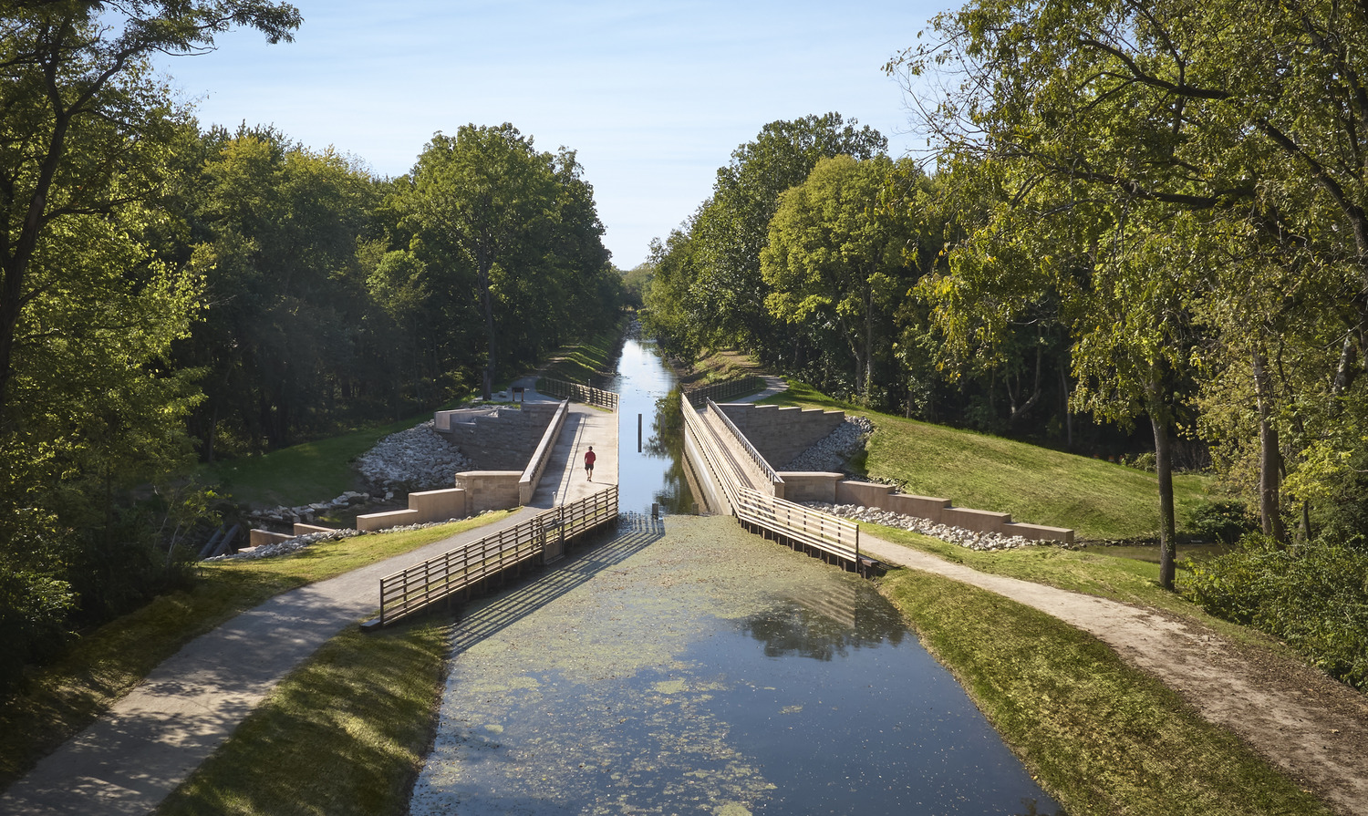 A narrow canal with a water lock surrounded by green trees and grassy paths on both sides under a clear blue sky.