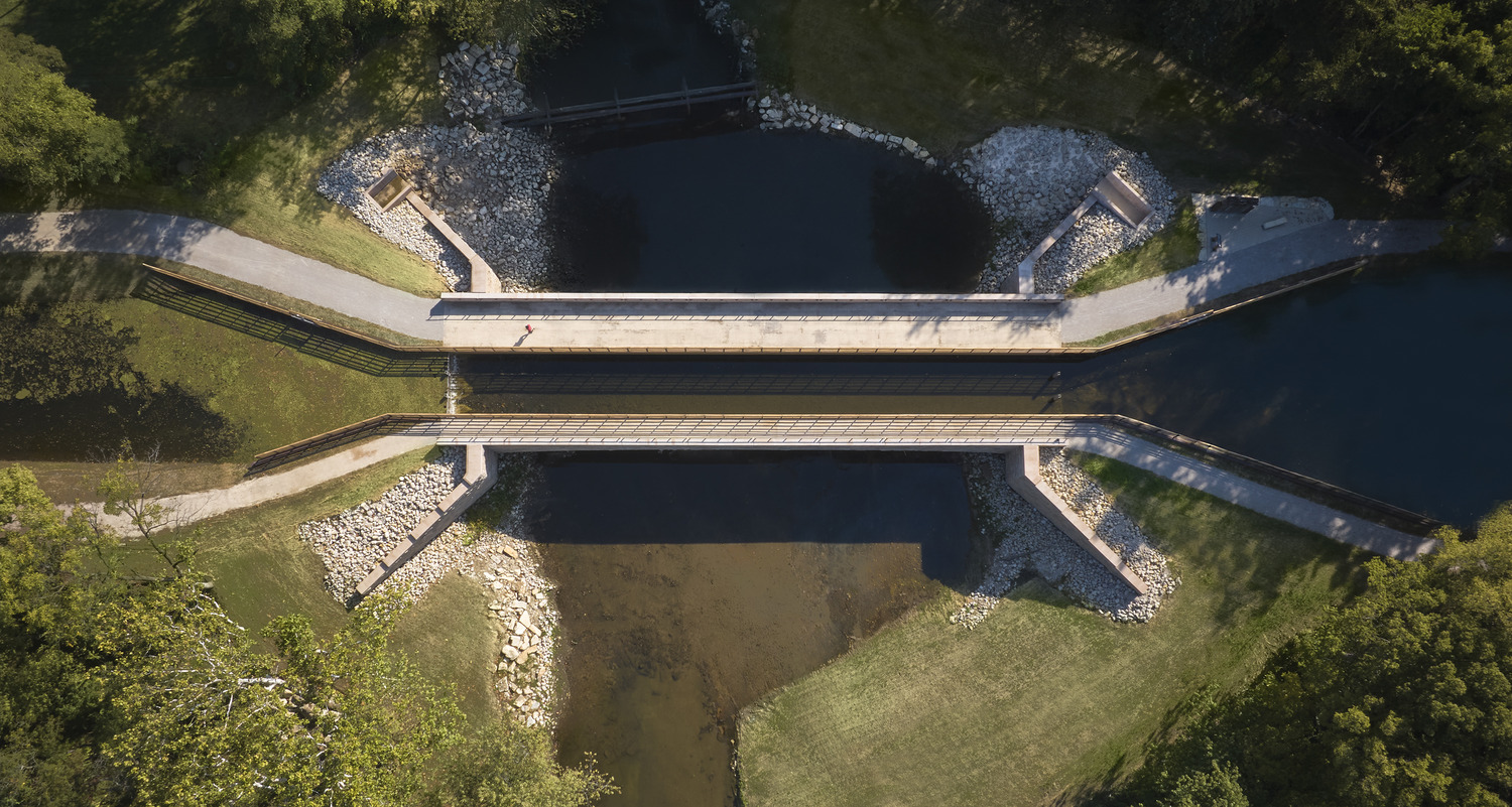 Aerial view of a bridge crossing over a waterway with pathways on both sides, surrounded by grass and trees.