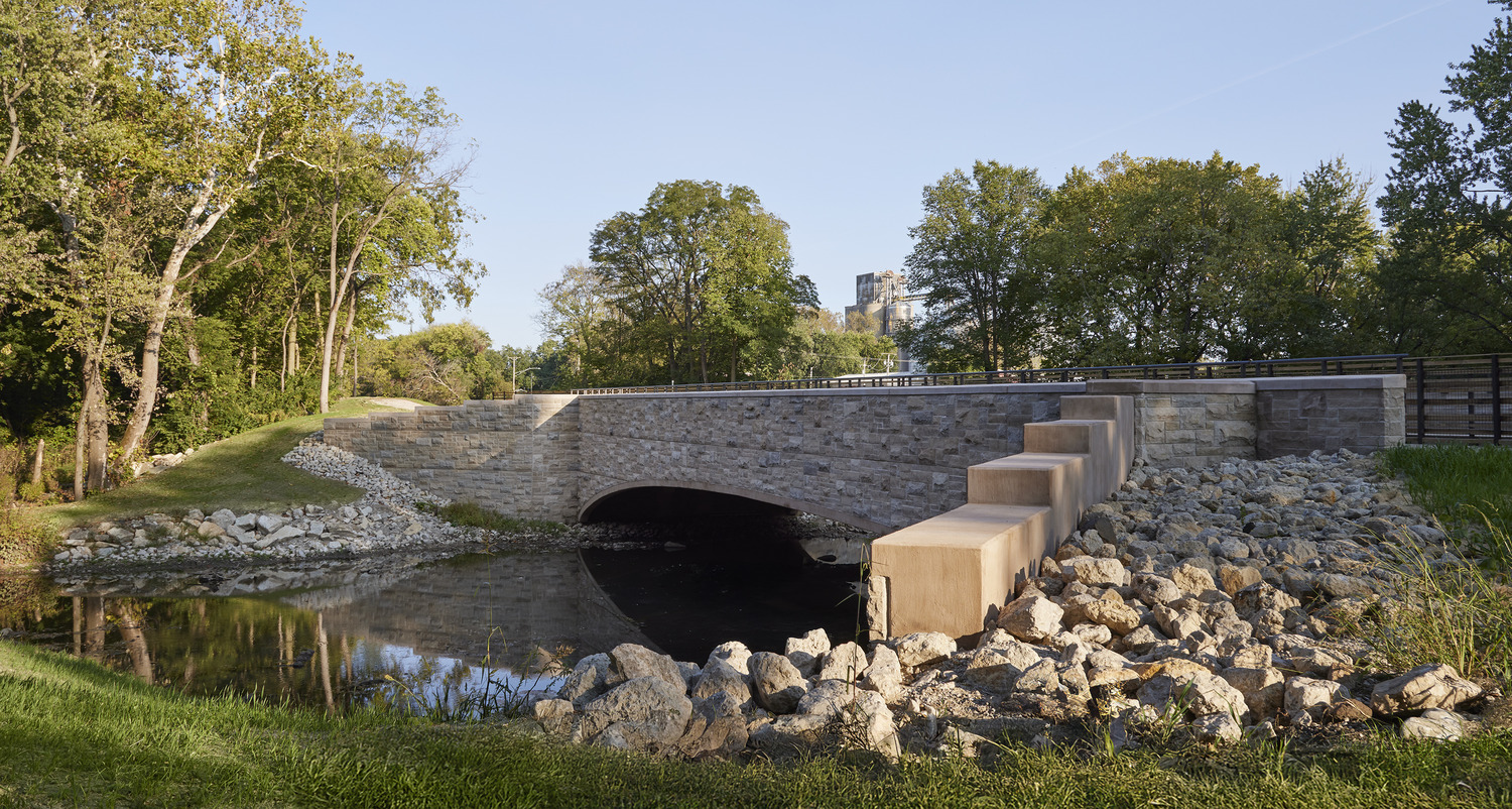 A stone arch bridge crosses over a small creek, surrounded by rocks and trees, with a building visible in the background.