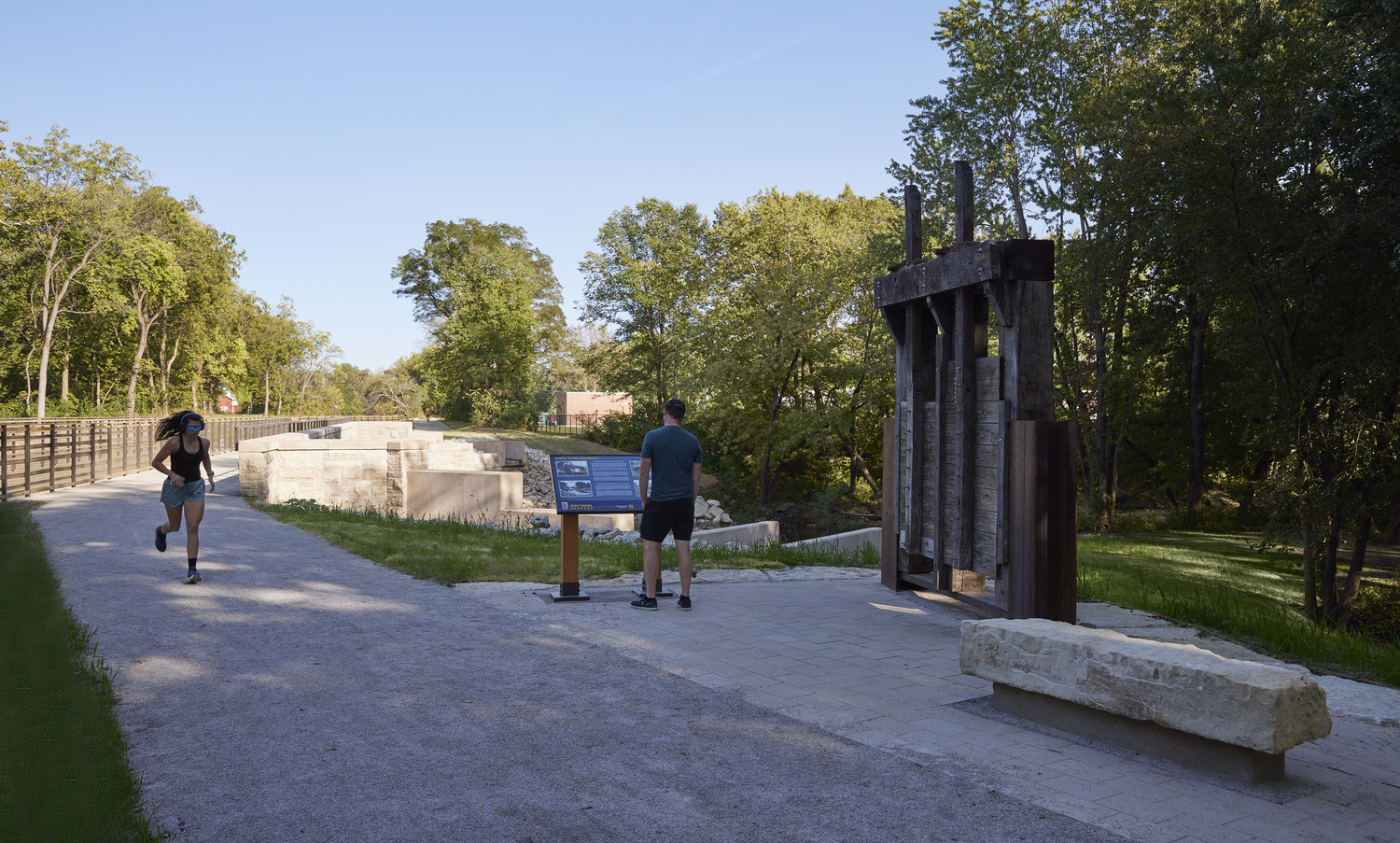 Two people are on a gravel path by an informational sign and a sculptural structure, surrounded by trees and greenery on a sunny day.