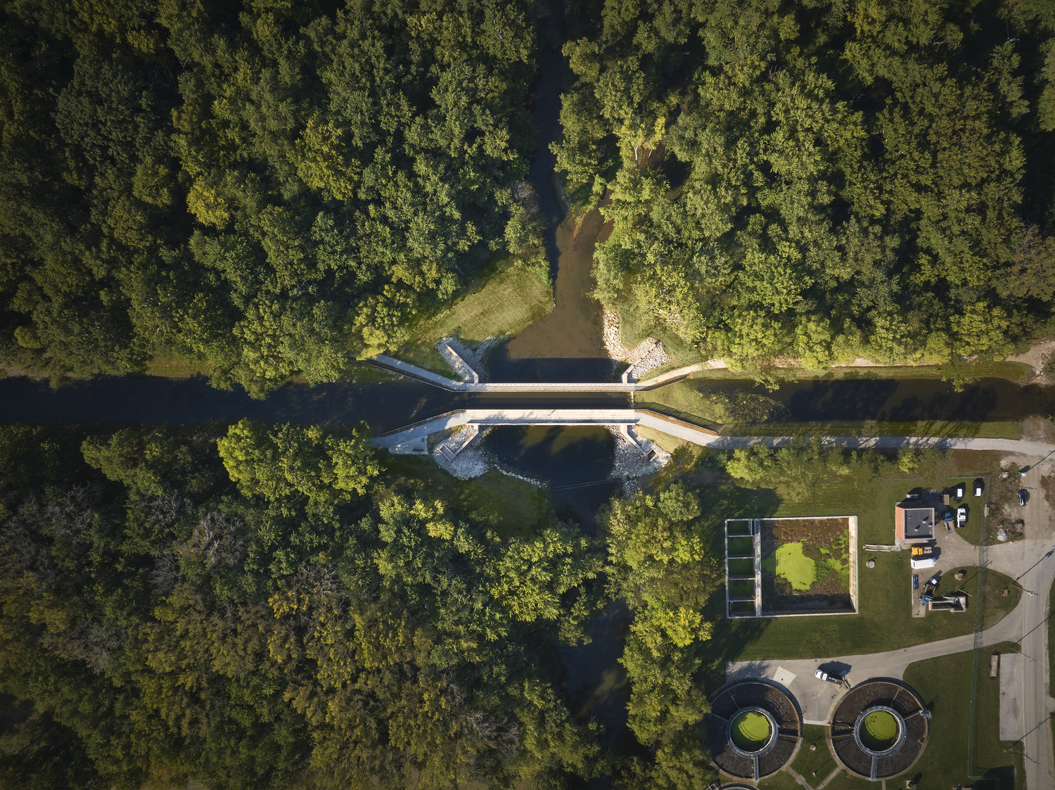 Aerial view of a small bridge crossing a narrow river surrounded by dense trees, with adjacent buildings and circular structures on the lower right.