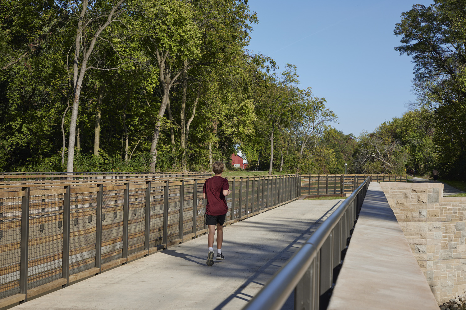 A person in a maroon shirt jogs alone on a paved bridge surrounded by trees under a clear blue sky.