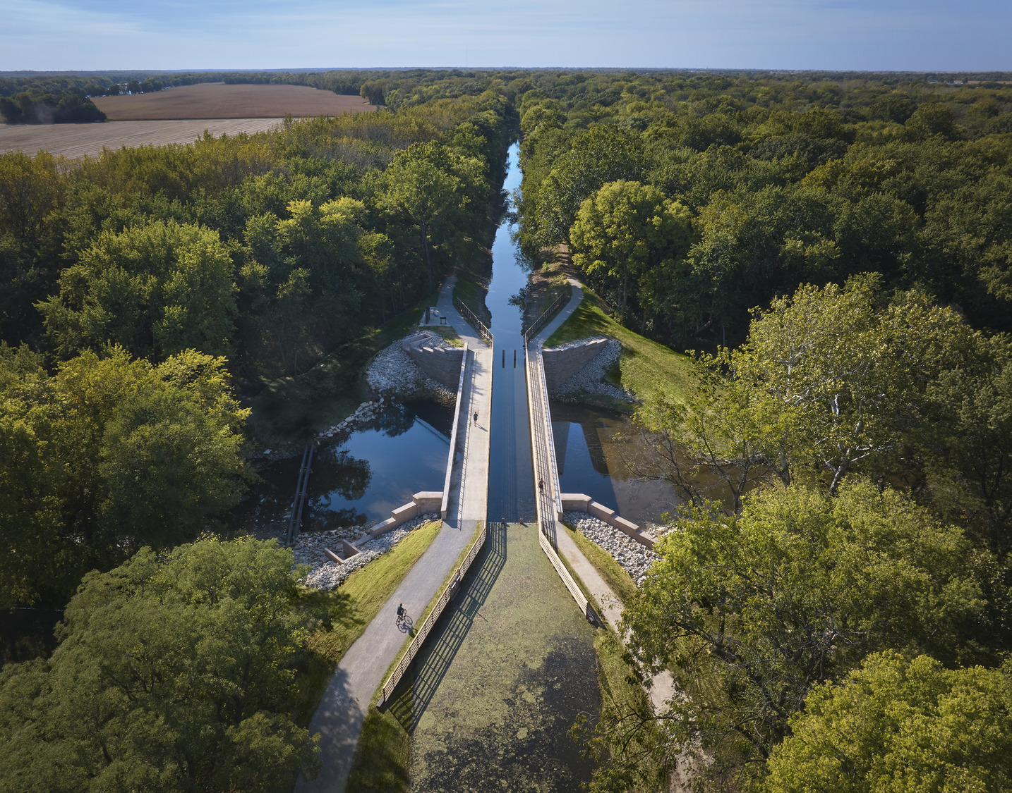 Aerial view of a bridge crossing over a canal surrounded by dense green trees and fields under a clear sky, with a few people visible on the bridge and nearby paths.