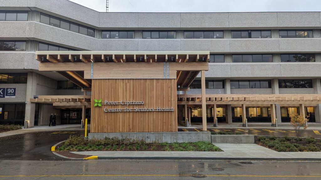 The exterior of a multi-story building with the sign "Peter C. Connell Centre for Seniors Health" on a wooden facade, with a covered entrance and visible parking spaces.
