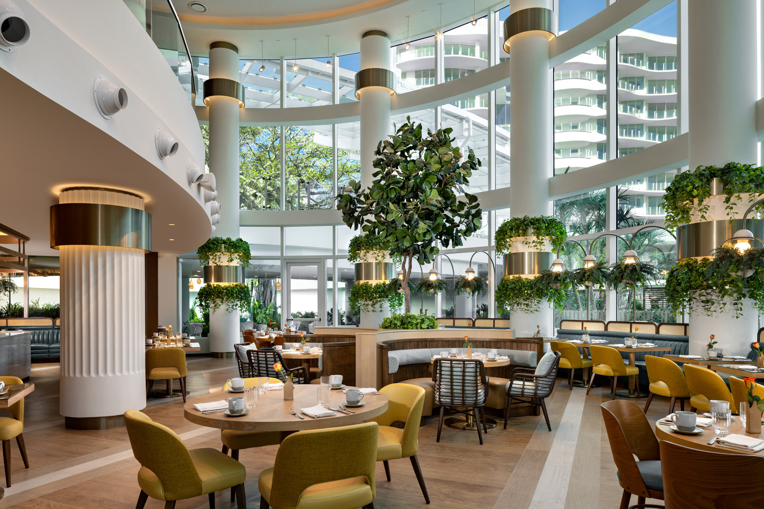 Intérieur d'un restaurant moderne avec de grandes fenêtres, de hautes colonnes blanches, des plantes vertes, des chaises jaunes et des tables de salle à manger, le tout baigné de lumière naturelle.