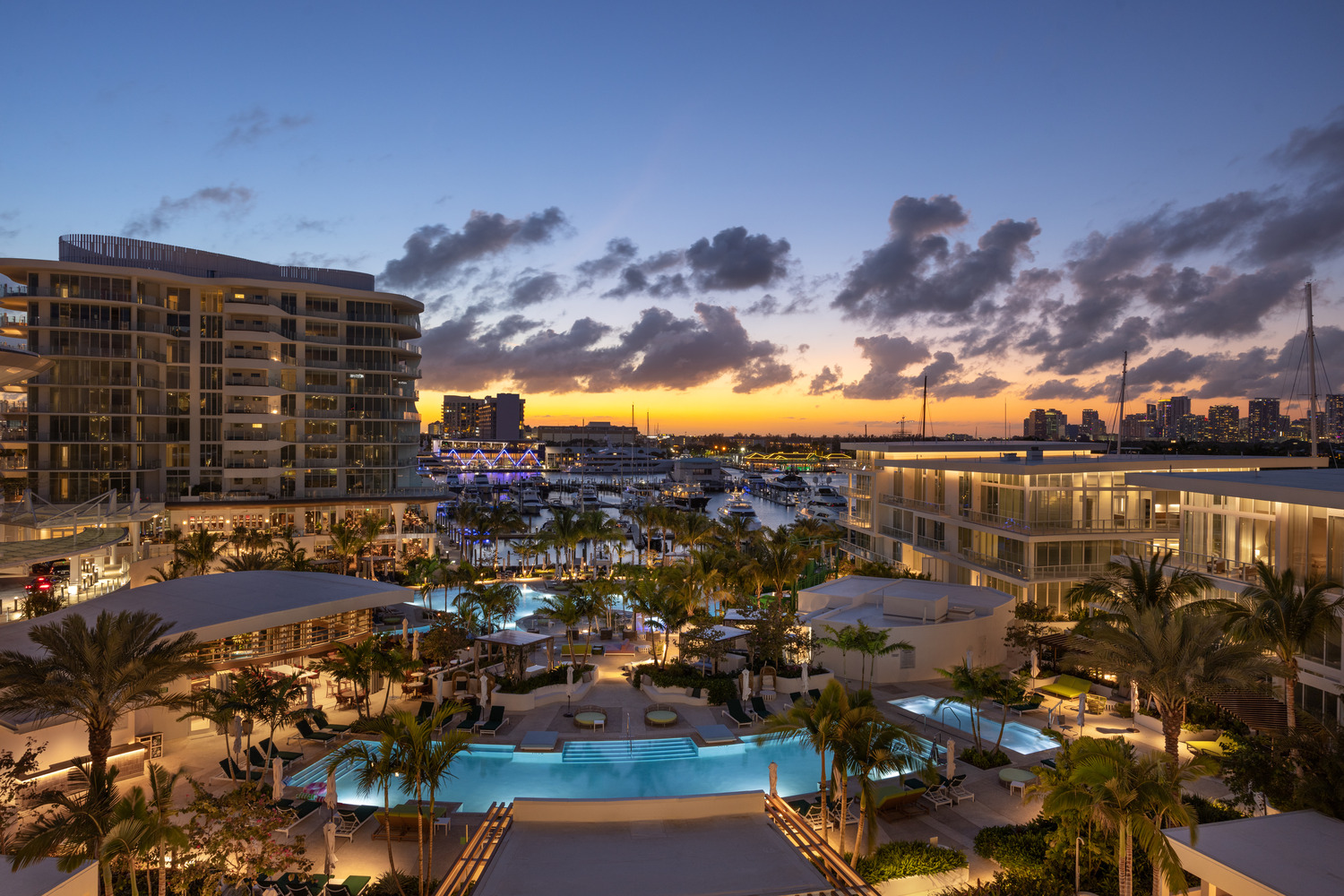 Une station balnéaire avec des piscines, des palmiers et des yachts est montrée au coucher du soleil, avec des bâtiments modernes et la ligne d'horizon d'une ville en arrière-plan.