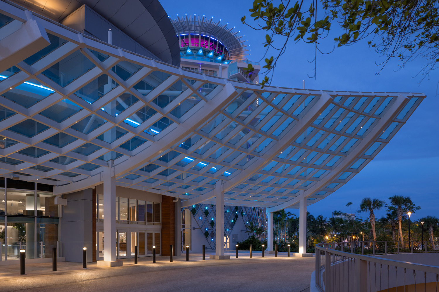 Un bâtiment moderne avec une verrière géométrique et des lumières bleues, photographié au crépuscule avec des palmiers visibles en arrière-plan.