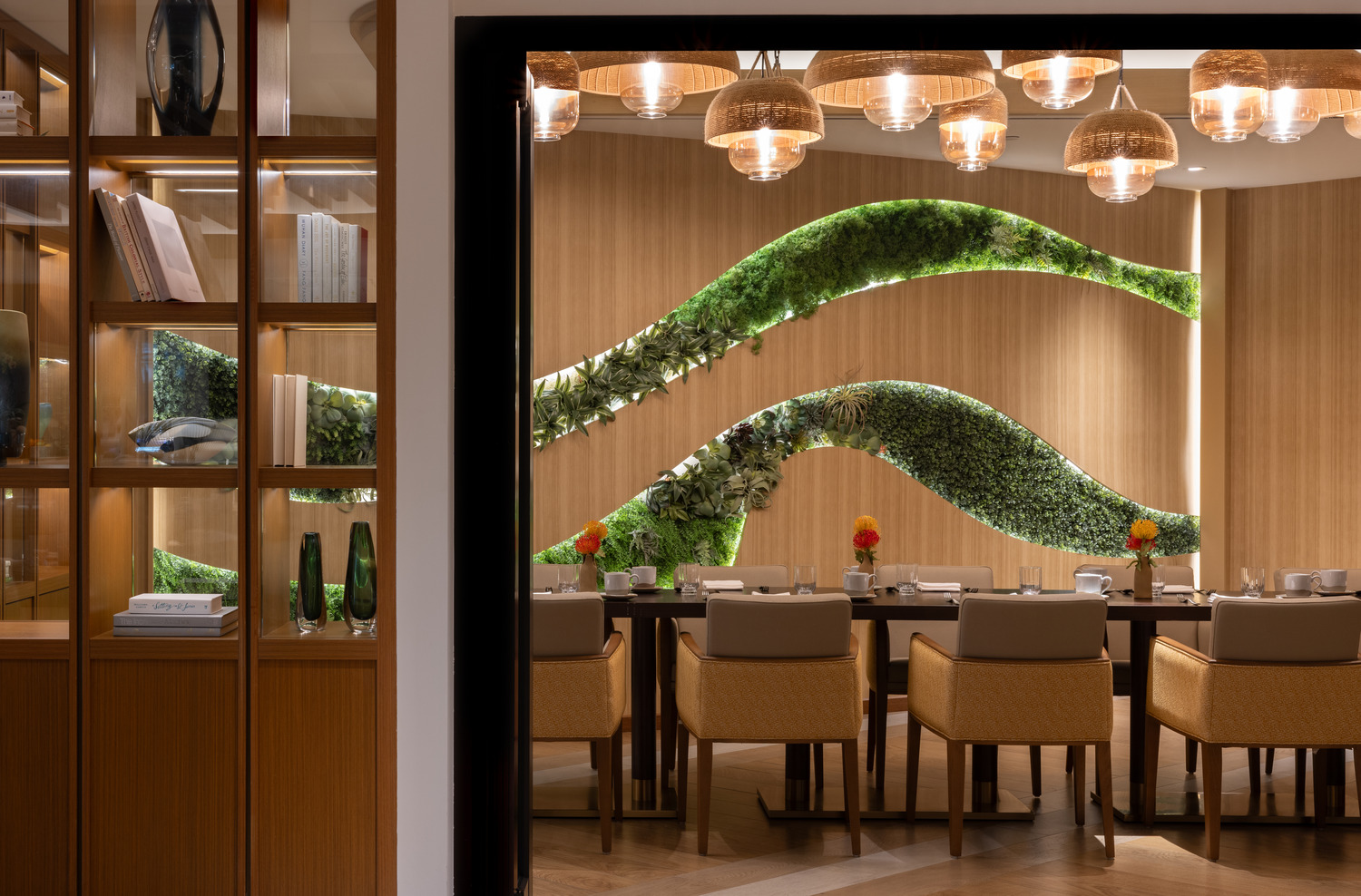 Une salle à manger moderne avec une longue table, des chaises beiges, des accents muraux verts, des panneaux en bois, des lampes suspendues et des étagères encastrées présentant des objets décoratifs.