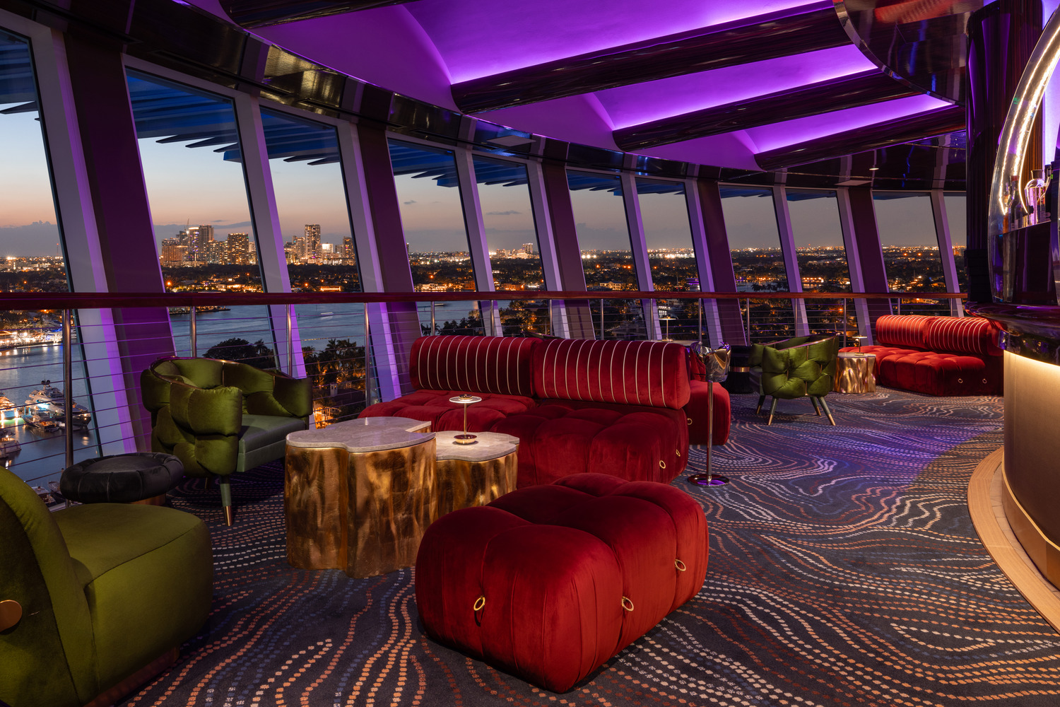Salon moderne avec des sièges rouges et verts en peluche, des tables rehaussées d'or et des vues panoramiques de la ville à travers de grandes fenêtres au coucher du soleil, sous des plafonniers violets.