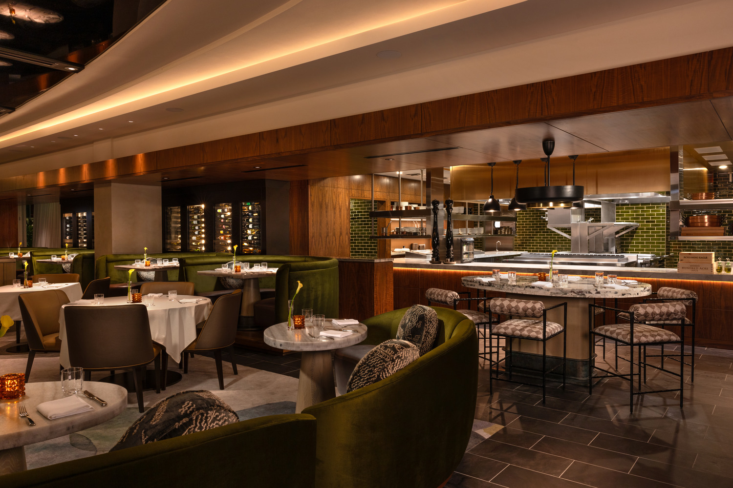 Intérieur d'un restaurant moderne avec des cabines en velours vert, des tables rondes, une cuisine ouverte et un éclairage d'ambiance chaleureux.