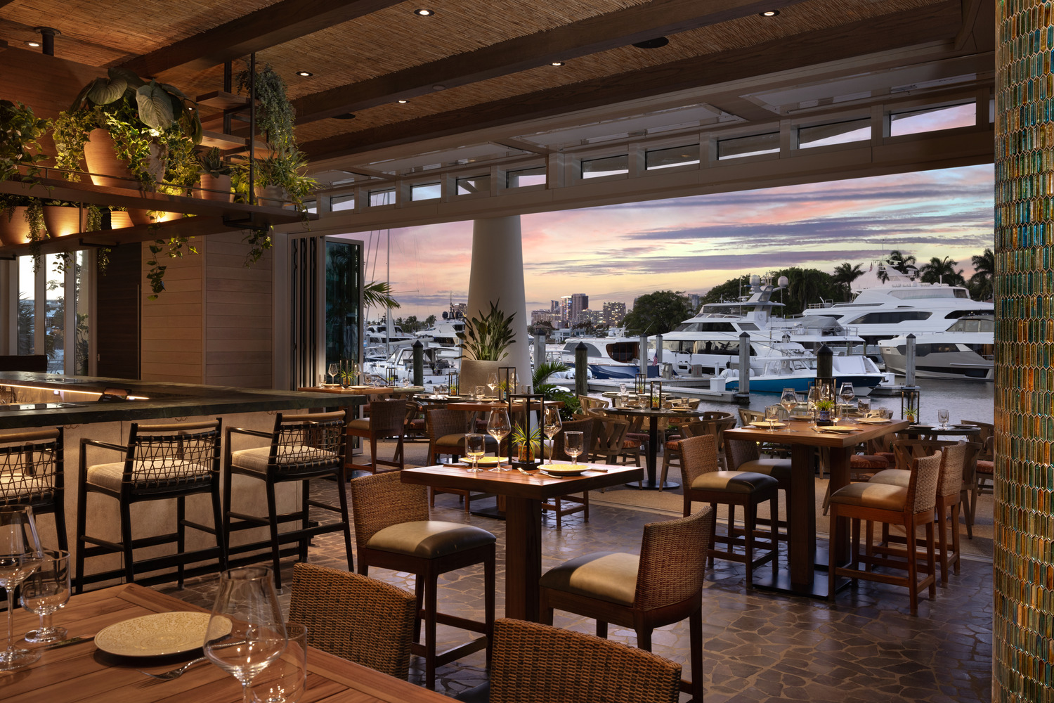 Restaurant haut de gamme avec mobilier en bois et sièges de bar, situé à côté de grandes fenêtres donnant sur les yachts et le port de plaisance au coucher du soleil.