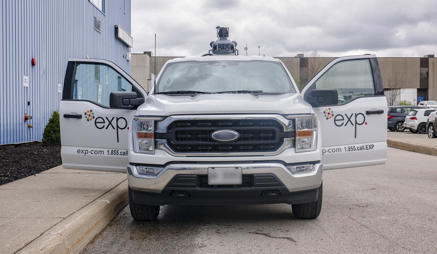 A white pickup truck with both front doors open, featuring "exp." branding and a roof-mounted sensor, is parked on a street near industrial buildings.