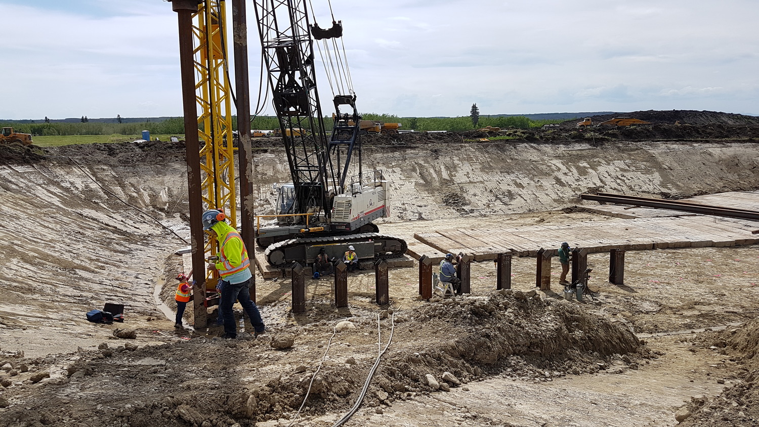 Construction workers install steel piles at a large excavation site, with a crane and equipment visible in the background under a partly cloudy sky.