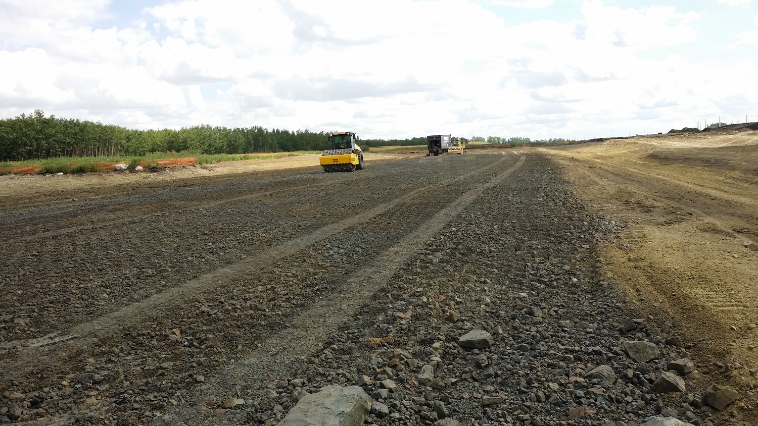 A construction site with a gravel road under development, featuring a yellow road roller and another vehicle working in the background.