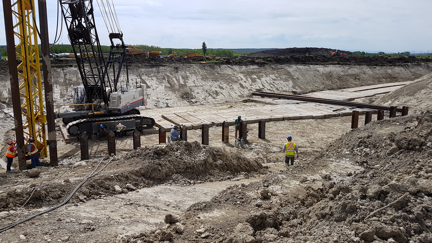 Construction workers at a large excavation site operate machinery and install vertical steel beams along the edge of an earthen pit.
