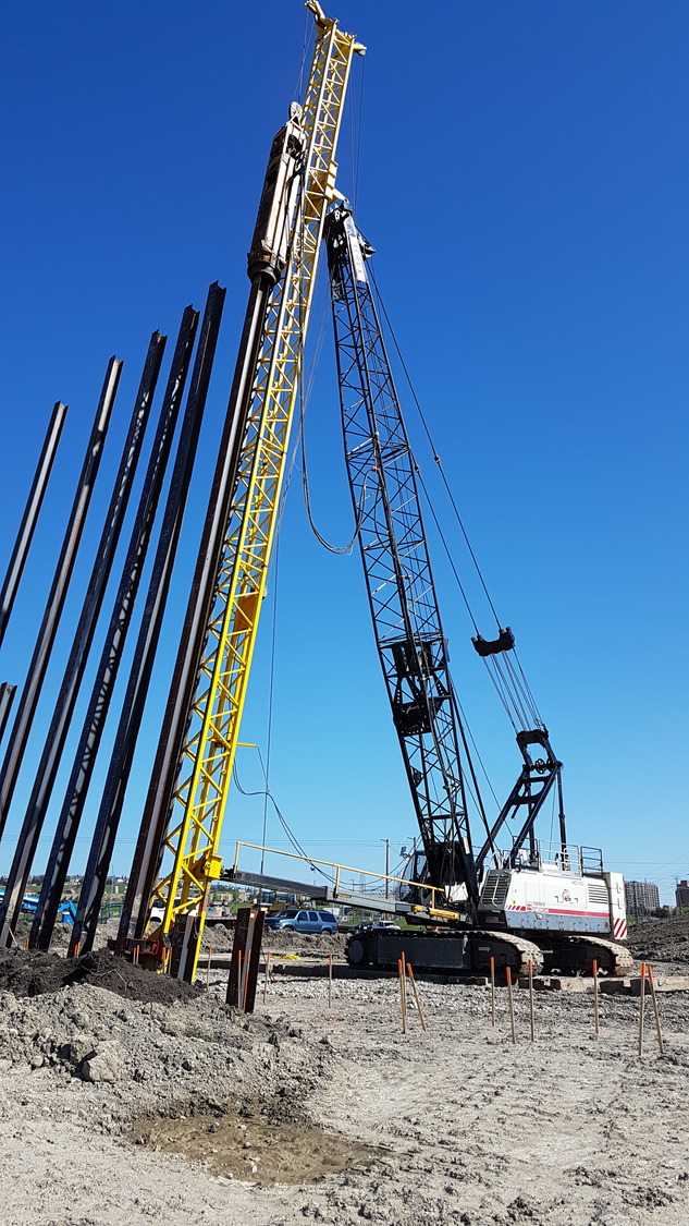 A construction site with a large crane and a yellow pile driver installing vertical steel beams into the ground under a clear blue sky.