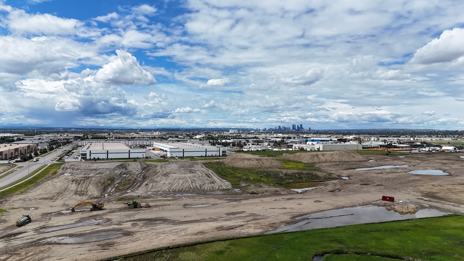 A large, muddy construction site with machinery in the foreground, industrial buildings in the midground, and a city skyline in the distance under a partly cloudy sky.