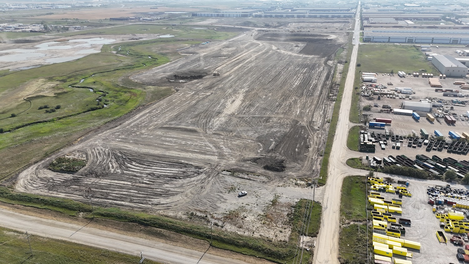 Aerial view of a large, cleared construction site with dirt paths, surrounded by industrial buildings, vehicles, and grassy areas.