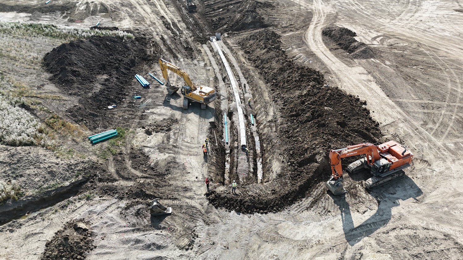 Aerial view of two excavators and workers installing pipes in a long trench at a large construction site.