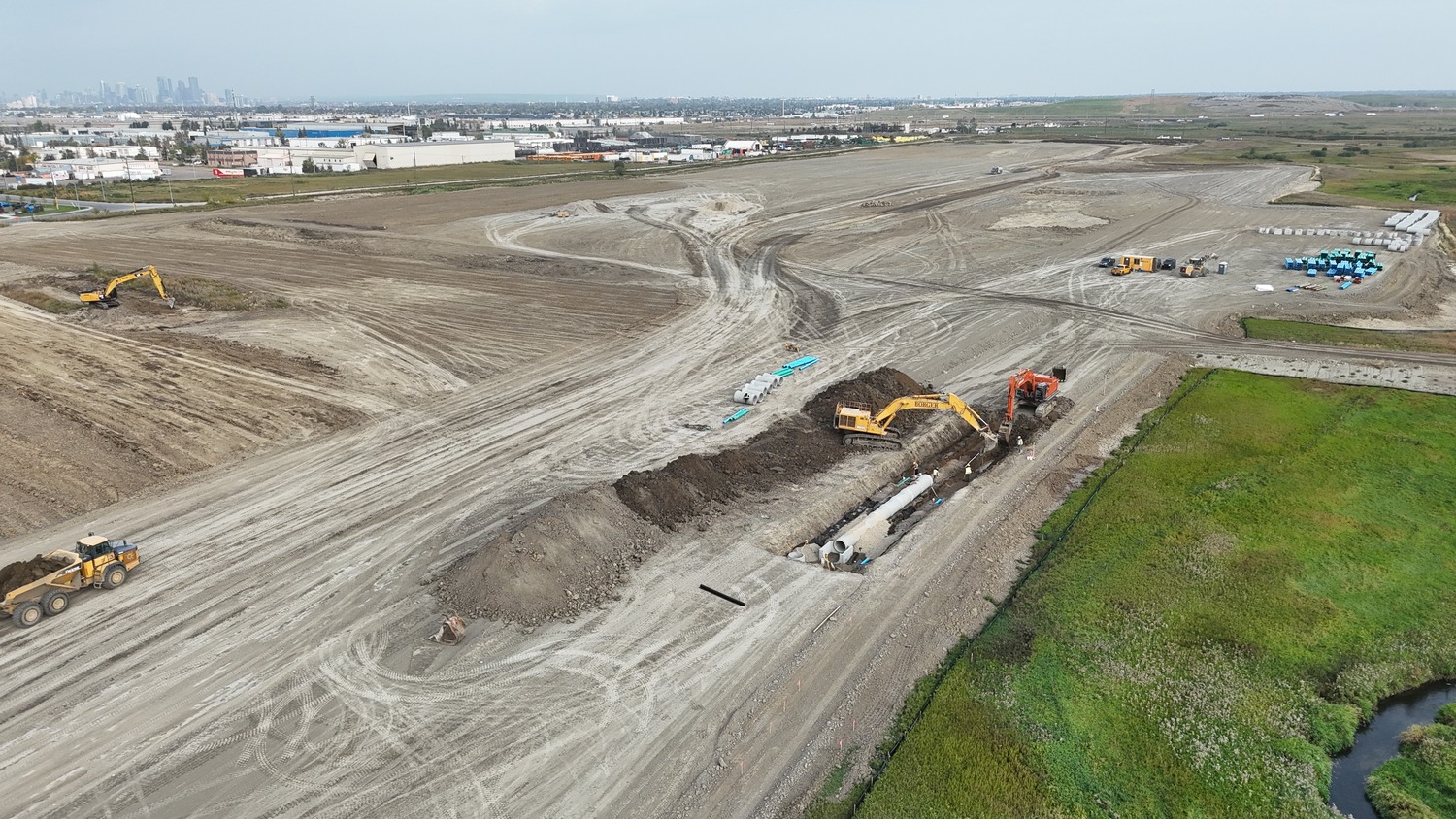 Aerial view of a construction site with excavators, pipes, and dirt piles; industrial buildings and city skyline visible in the background.
