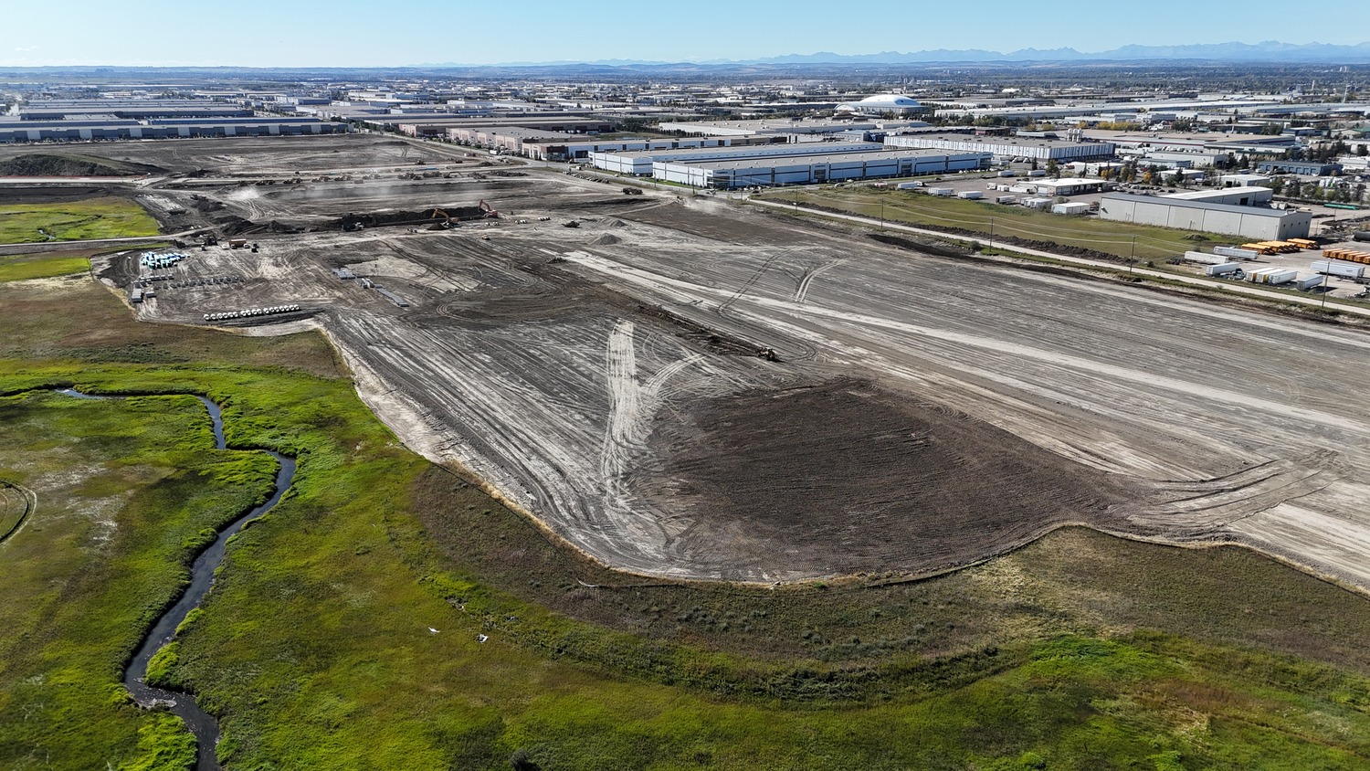 Aerial view of a large construction site with leveled dirt, heavy machinery, and surrounding industrial buildings; grassy area with a stream in the foreground.