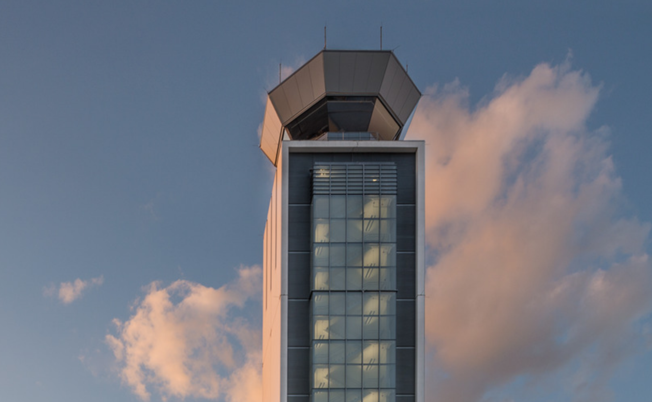 A modern air traffic control tower stands against a blue sky with scattered clouds in the background.