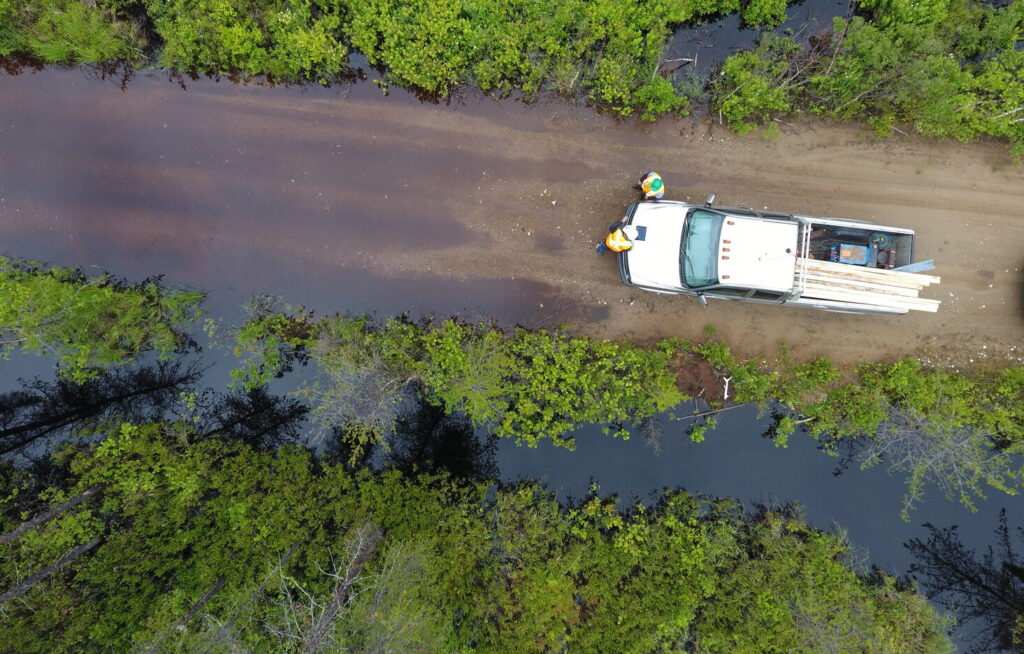 Aerial view of a truck carrying lumber parked on a dirt road surrounded by water and dense green foliage, with two people standing beside it.