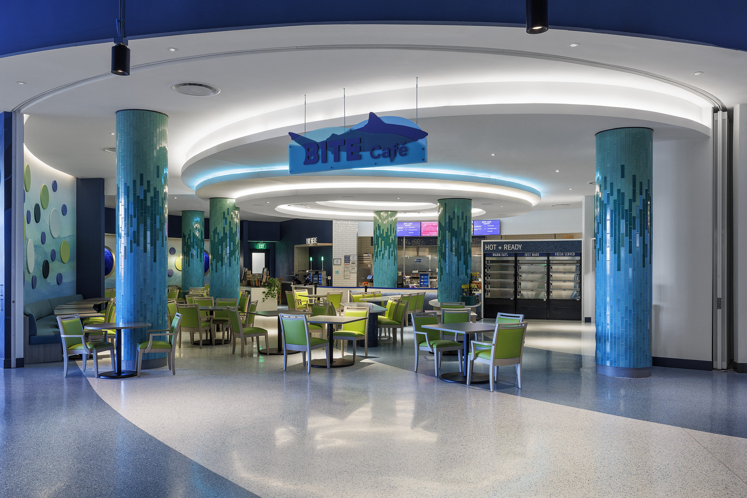 A modern café with green and blue décor, round tables with chairs, turquoise pillars, and a 