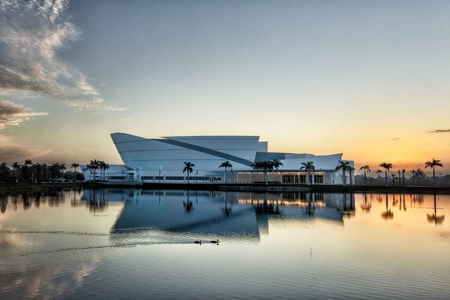 A modern white building with curved lines is reflected in a lake at sunset, surrounded by palm trees and a clear sky.