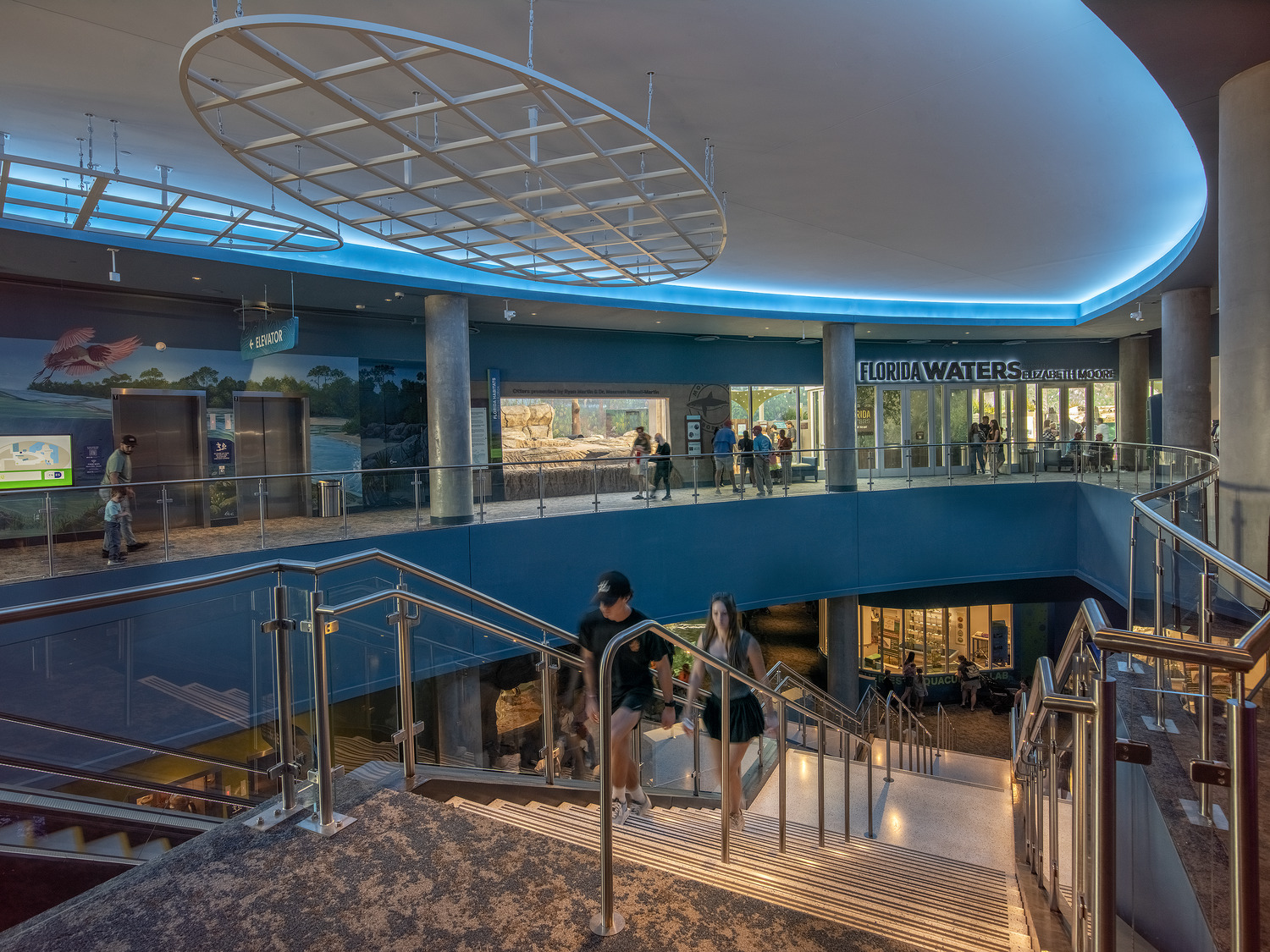 Wide staircase inside a modern building with people walking; blue accent lighting above; sign reads 