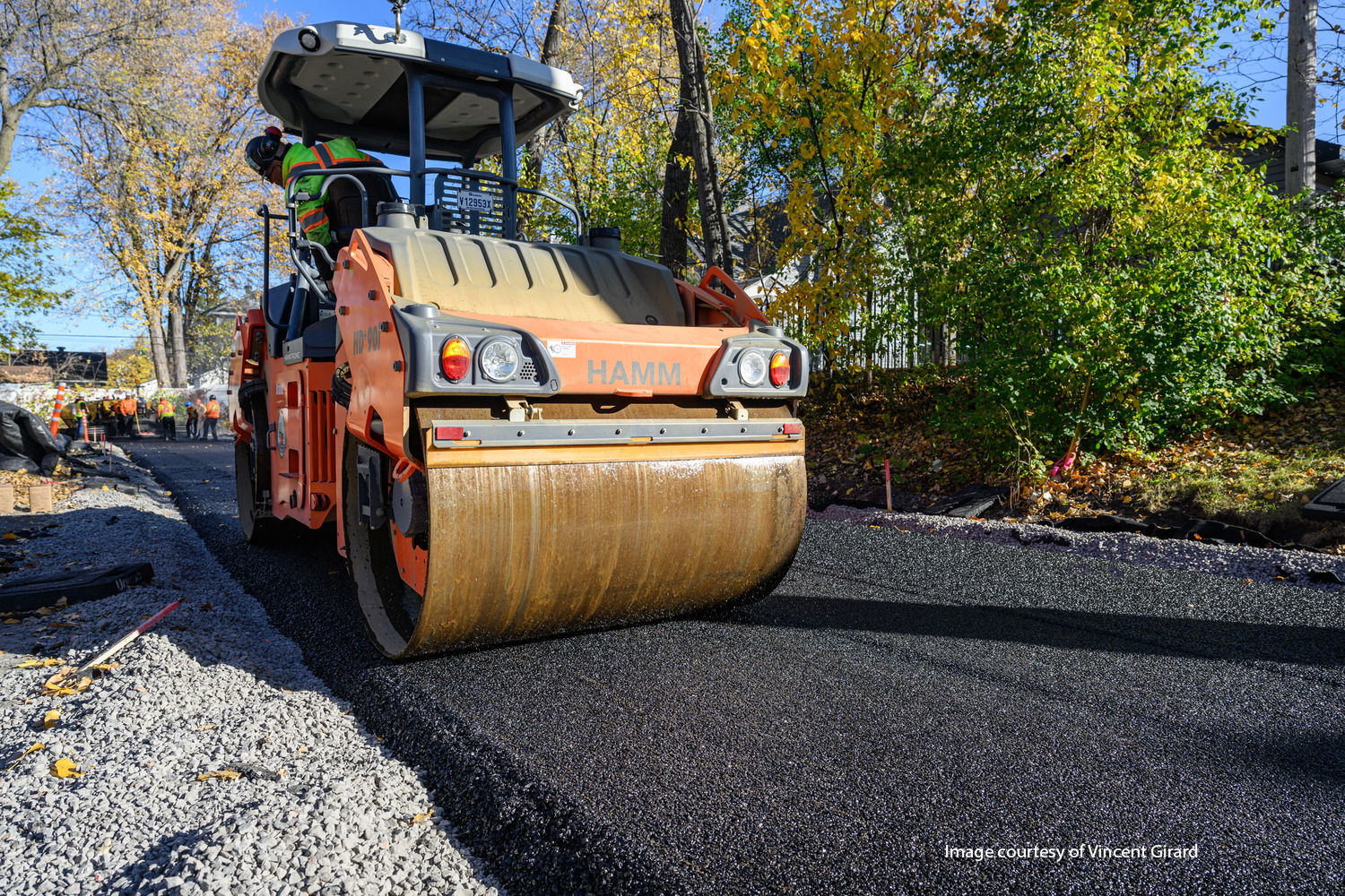 A road roller compacts freshly laid asphalt on a road surrounded by trees during daytime.