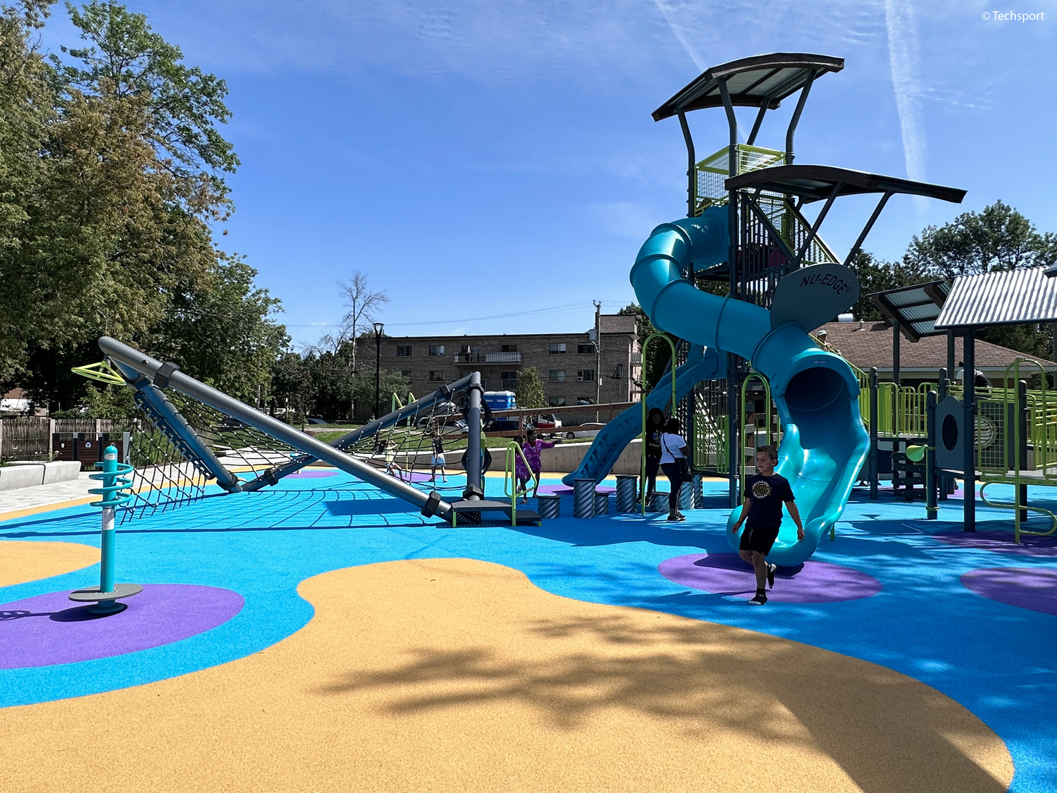 Children play on a modern playground featuring slides, climbing structures, and a bright blue and yellow rubber surface on a sunny day.