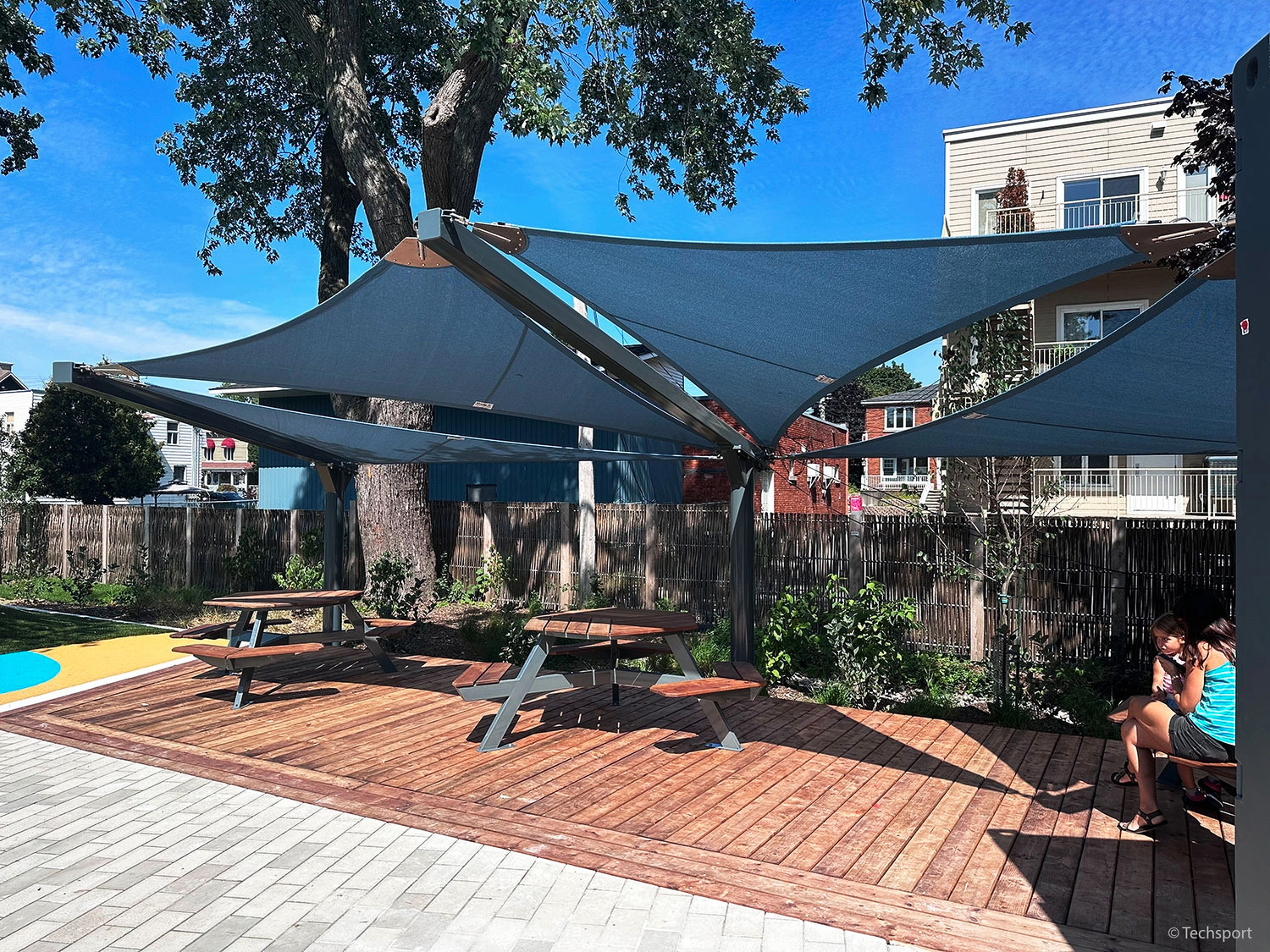 Two picnic tables sit under large, modern shade structures on a wooden deck in an outdoor park area, with trees and buildings in the background.
