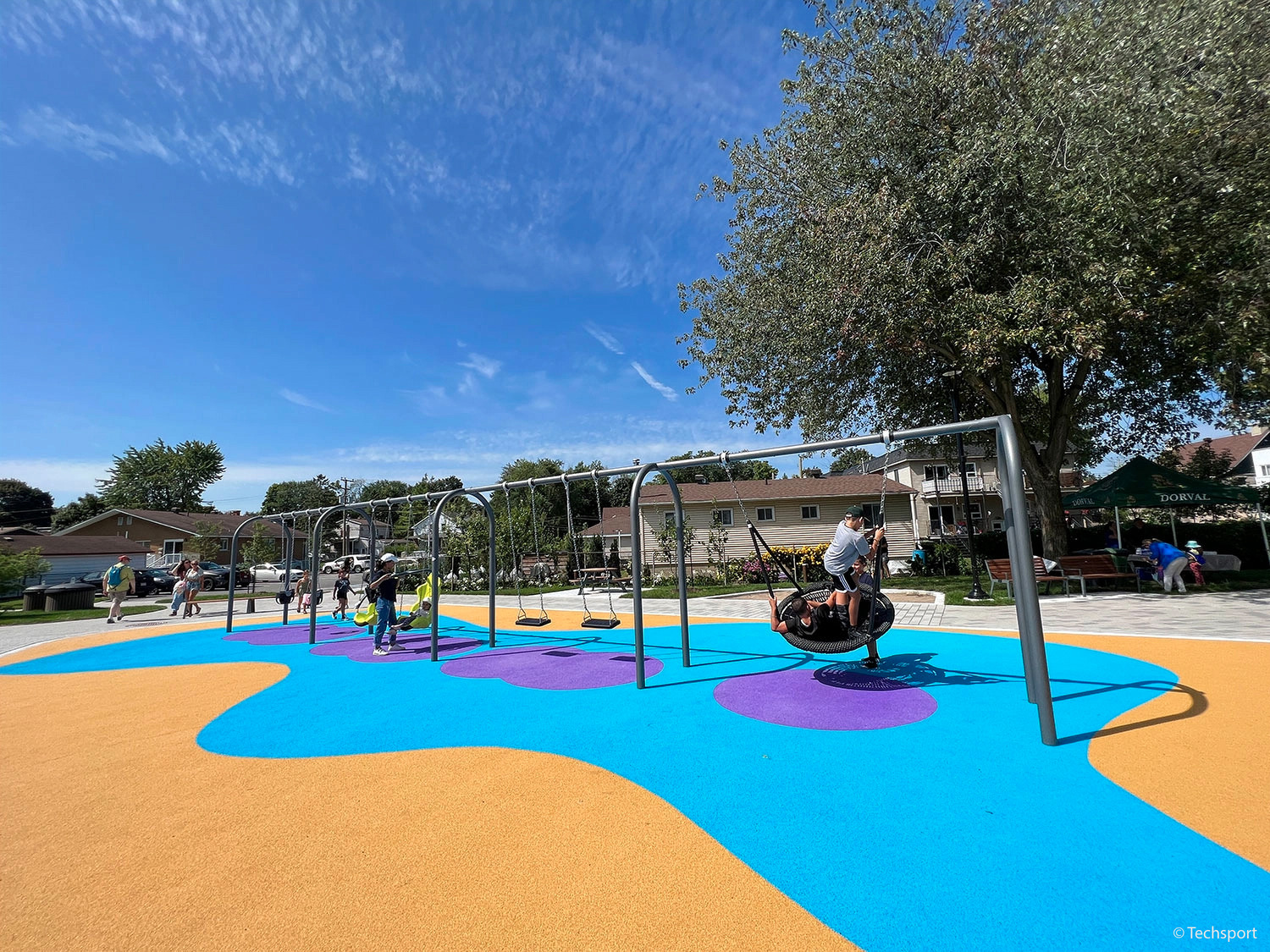 Children and adults are using swings on a modern playground with bright blue and orange rubber flooring under a clear sky.