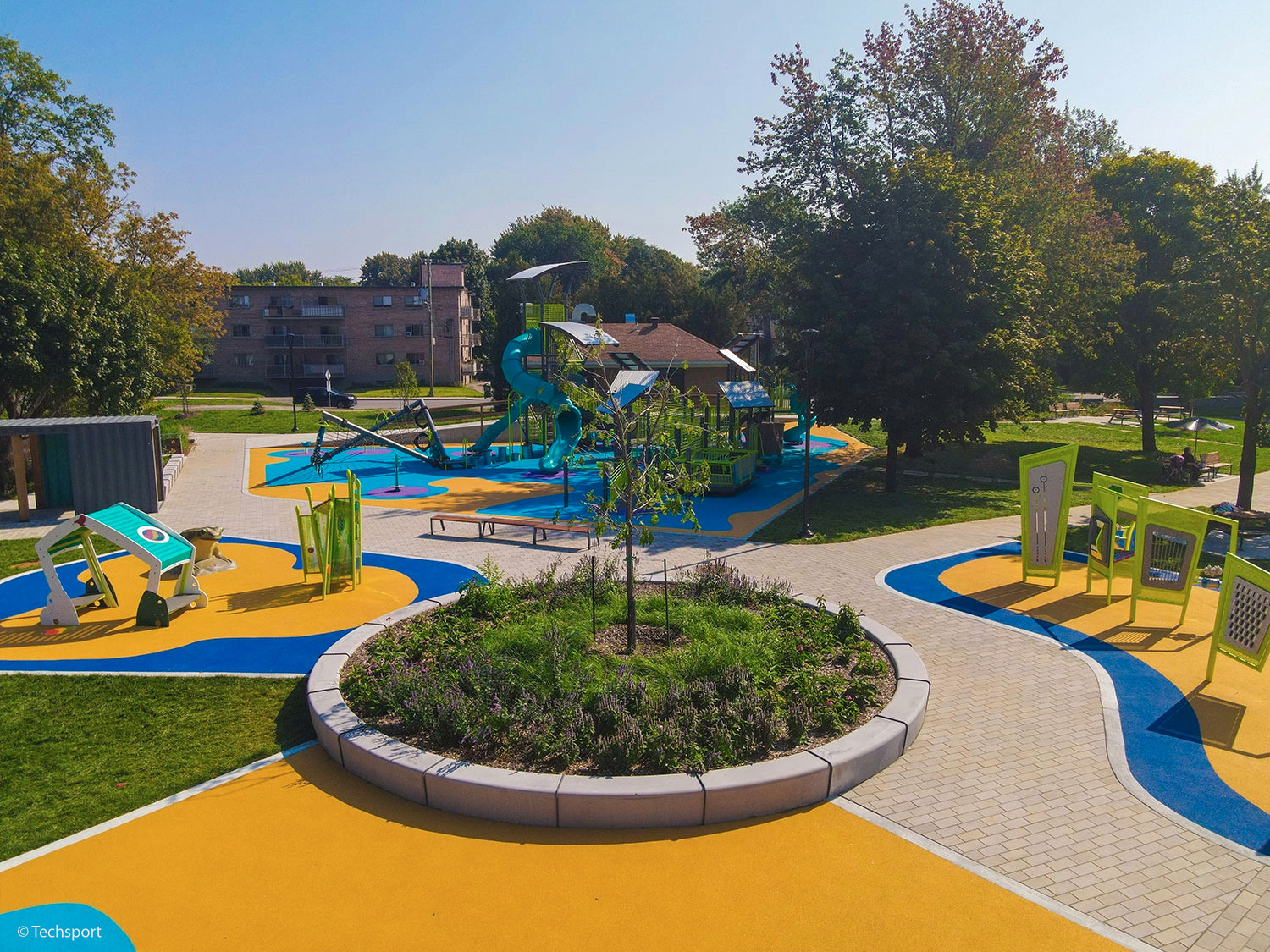 A modern outdoor playground features colorful play equipment, winding paths, and green landscaping, with trees and apartment buildings in the background.