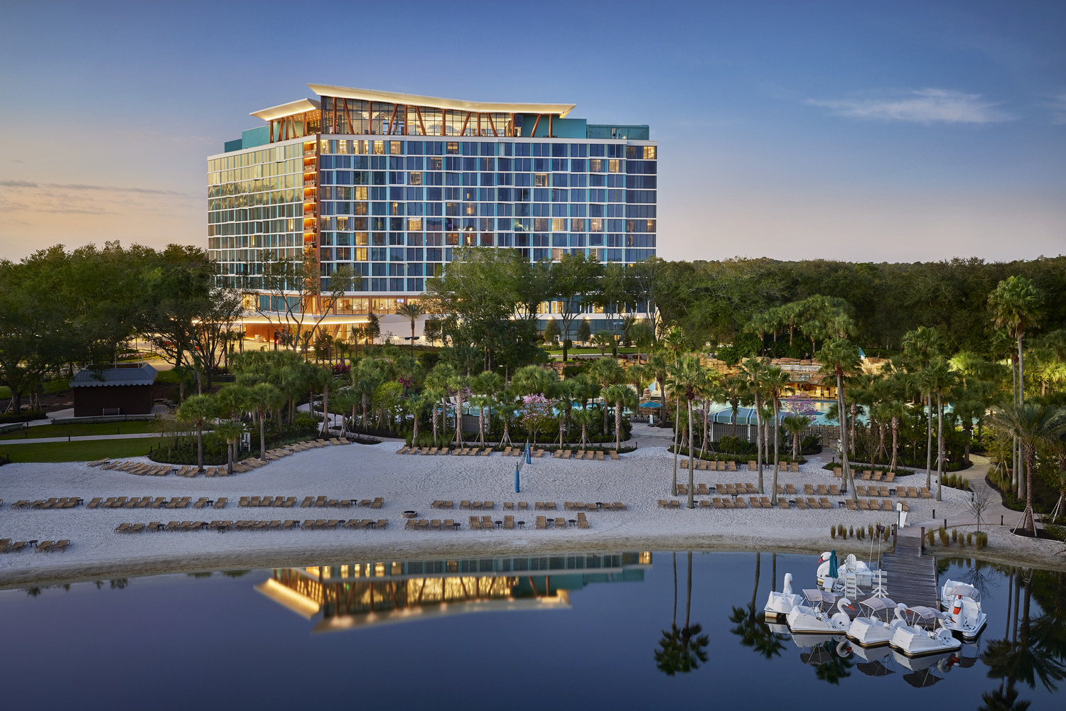 A modern hotel building, the Disney Swan Reserve, with a glass exterior overlooks a sandy beach area, palm trees, lounge chairs, and calm water with pedal boats in the foreground.