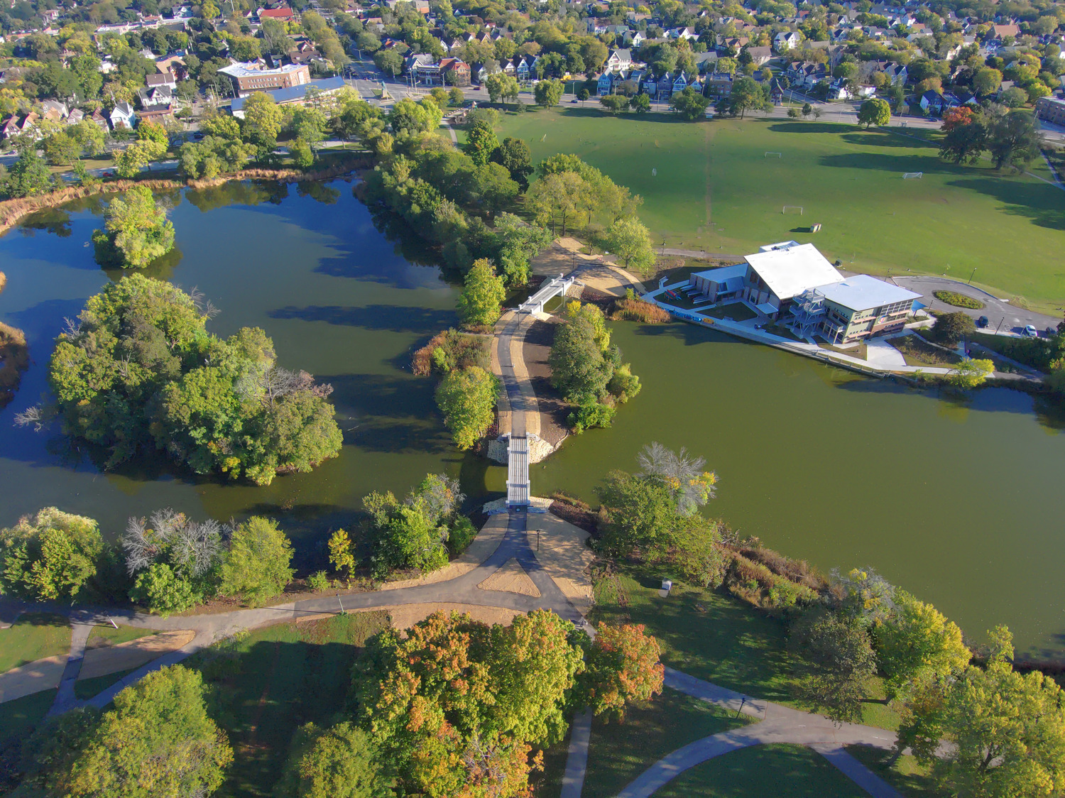 Aerial view of a park with two ponds, a footbridge connecting walking paths, open green fields, and a modern building near the water.