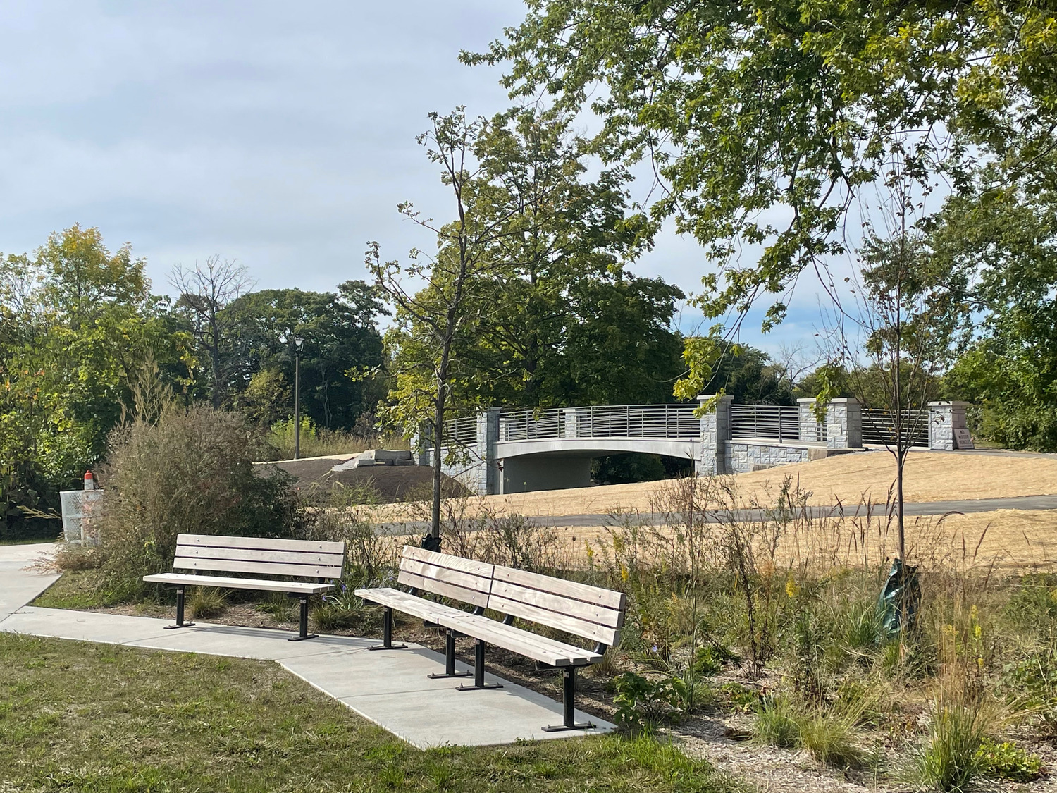 Two empty wooden benches face a grassy park area with a modern white pedestrian bridge in the background, surrounded by trees and shrubs under a partly cloudy sky.