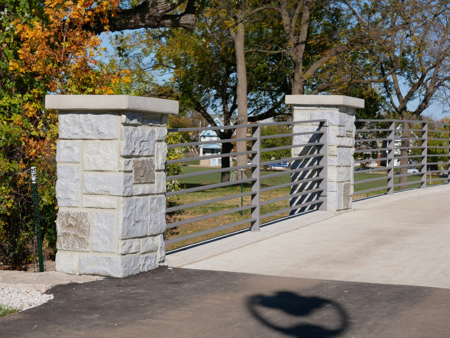 A paved path with stone pillars and metal railings, bordered by autumn trees and a shadow cast on the pavement in the foreground.