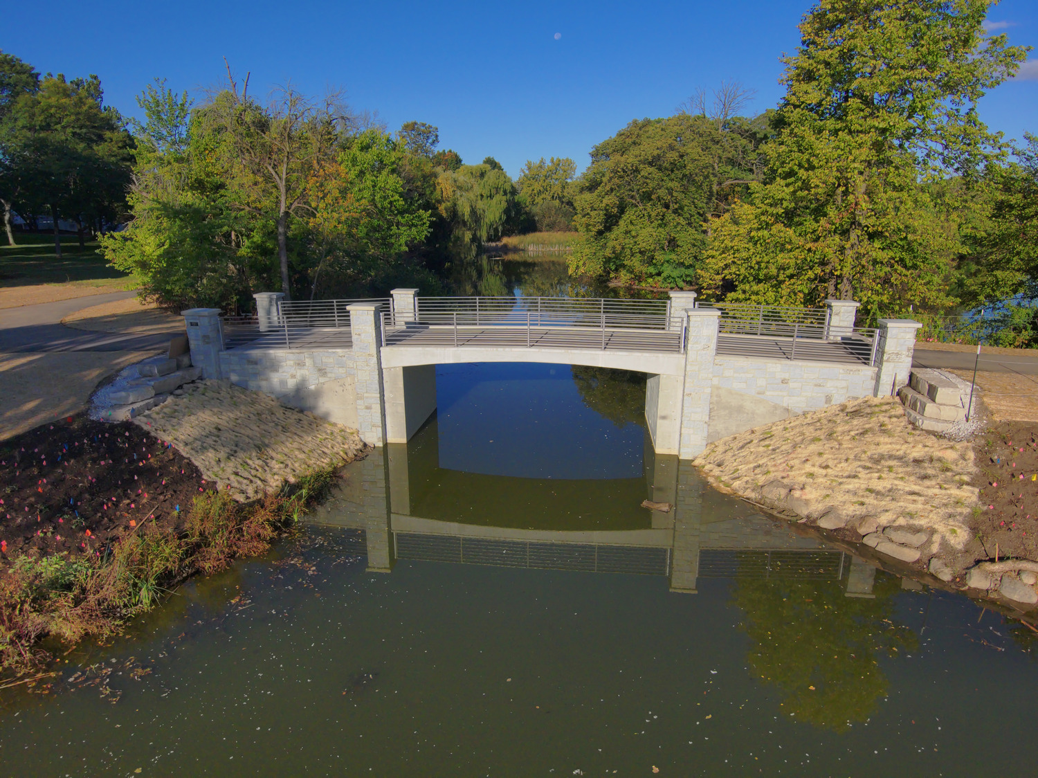 A small concrete and metal bridge crosses over a calm, narrow body of water, surrounded by trees and grassy banks under a clear blue sky.