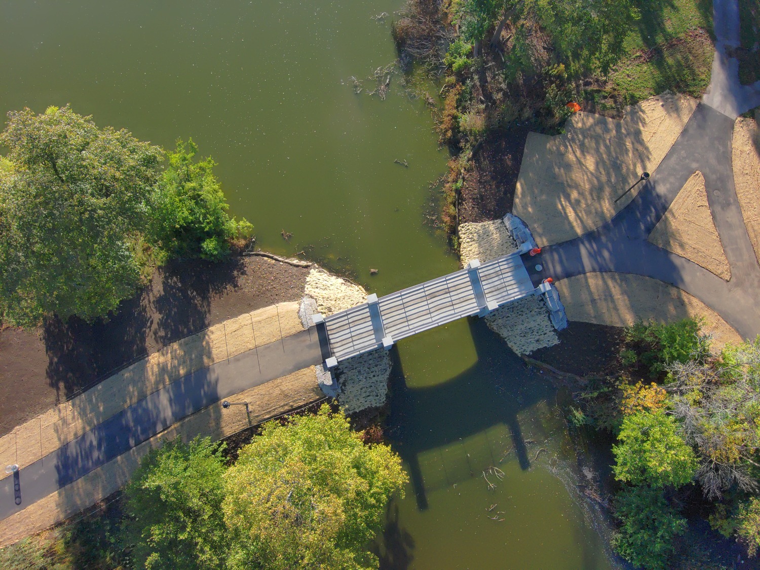 Aerial view of a small footbridge crossing a greenish pond, with paved walkways and surrounding trees casting shadows.