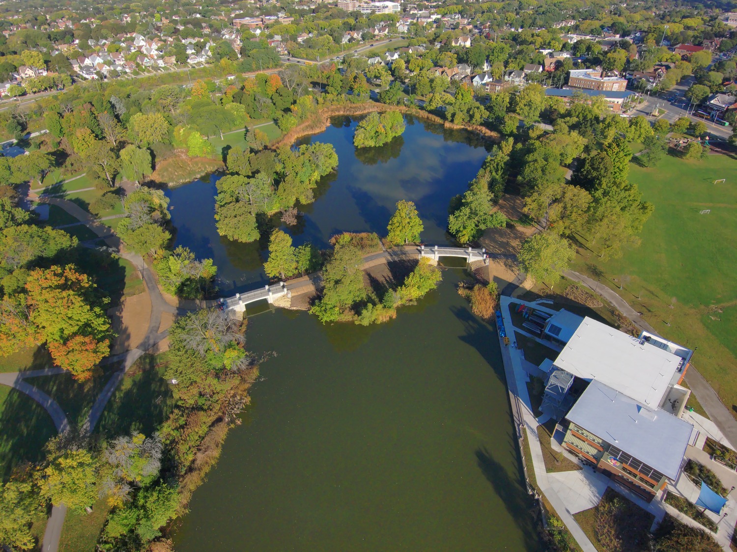 Aerial view of a park with two connected ponds, walking paths, green spaces, and a modern building near the water. Residential neighborhood in the background.