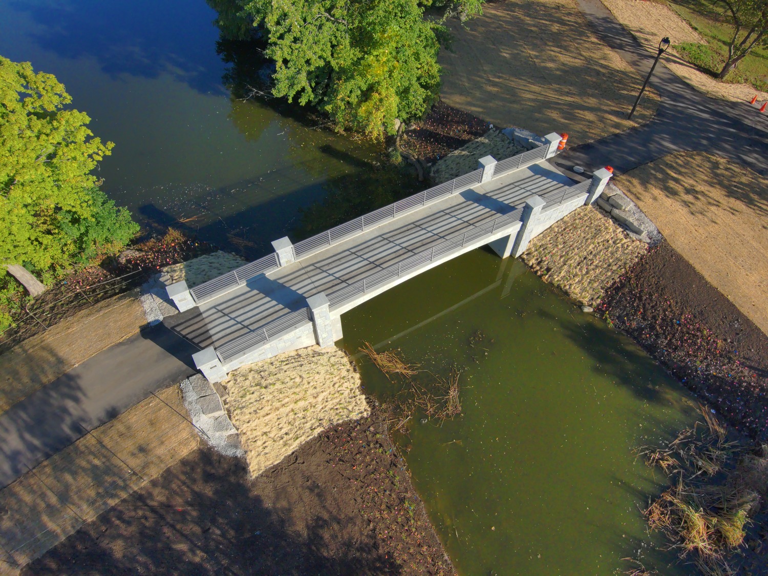 A small concrete pedestrian bridge crosses over a greenish pond, with trees, paths, and grassy areas on both sides.