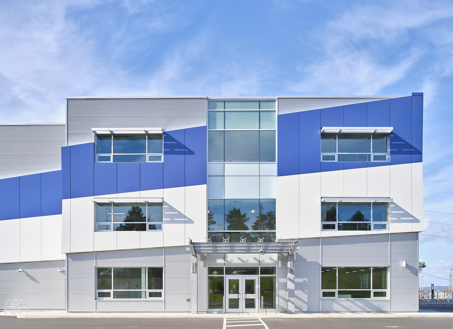 A modern, two-story building with blue and white exterior panels, large windows, and a glass entrance under a clear sky.